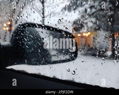 Primo piano delle gocce d'acqua sul finestrino laterale dell'auto durante le nevicate in inverno, contro lo sfondo dello specchio retrovisore e del Bokeh delle luci di strada. Foto Stock