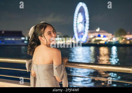 Felice bella giovane donna asiatica in abito sorridente e giro turistico illuminato ruota panoramica durante la crociera sul fiume di notte a Bangkok Foto Stock