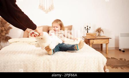 Il bambino non vuole condividere i giocattoli, grida isterico urlante. Una ragazza di due anni non dà un orso giocattolo. Papà prende un giocattolo morbido. Beige casa in Foto Stock