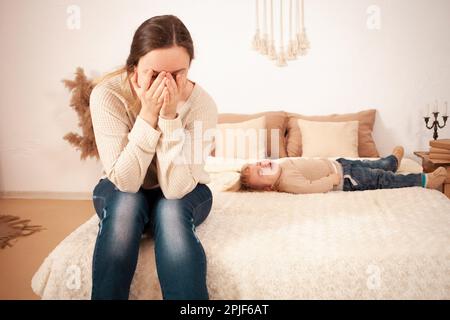 Mamma è stanca del bambino. Una bambina di due anni sdraiata sul letto urlando istericamente piangendo. La donna è infelice, copre il suo volto con le mani e lo fa Foto Stock