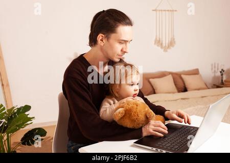 Un uomo lavora a casa con un notebook. Il bambino distrae dal lavoro, manca. Il padre e la figlia sono seduti al computer. Abbraccio di due anni Foto Stock