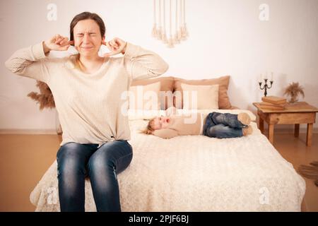 Mamma è stanca del bambino. Una bambina di due anni sdraiata sul letto urlando istericamente piangendo. La donna è infelice, copre il suo volto con le mani e lo fa Foto Stock