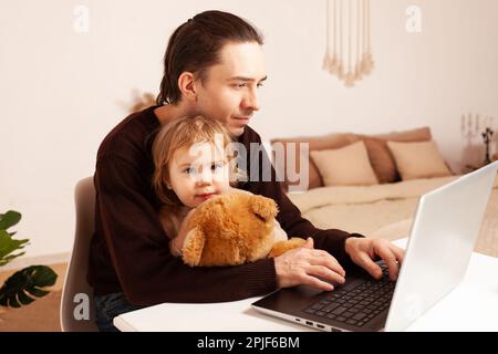 Un uomo lavora a casa con un notebook. Il bambino distrae dal lavoro, manca. Il padre e la figlia sono seduti al computer. Abbraccio di due anni Foto Stock