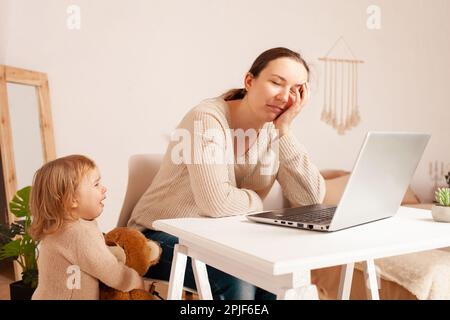 Una giovane madre in congedo di maternità si siede al computer portatile e lavora. Un bambino che urla istericamente grida distrae dal lavoro. Una donna è impegnata in freelancina Foto Stock