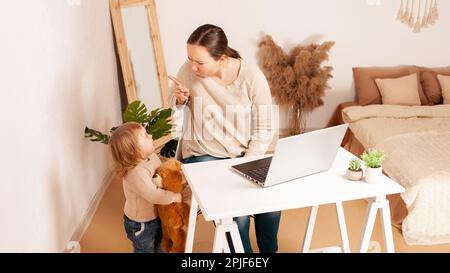 Una giovane madre in congedo di maternità si siede al computer portatile e lavora. Un bambino che urla istericamente grida distrae dal lavoro. Una donna è impegnata in freelancina Foto Stock