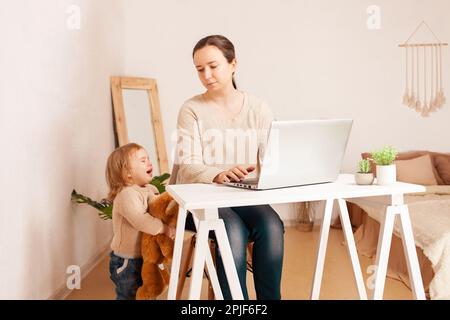 Una giovane madre in congedo di maternità si siede al computer portatile e lavora. Un bambino che urla istericamente grida distrae dal lavoro. Una donna è impegnata in freelancina Foto Stock