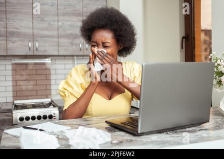 Una donna afro-americana malata starnutisce la tosse. Freelance, home Office, lavoro su notebook. Malsano Foto Stock