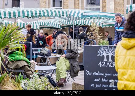 Giovane uomo con capelli lunghi e bearded seduto libro di lettura al caffè all'aperto al Frome Sunday Market, Somerset, Regno Unito il 2 aprile 2023 Foto Stock