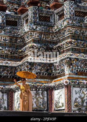Una graziosa signora Tailandese adornata in un vivace abbigliamento arancione tradizionale sorge orgogliosamente dal magnifico Tempio Wat Arun a Bangkok, in Thailandia, tenendo un ma Foto Stock