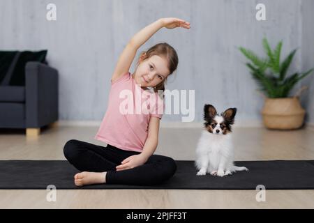 carina bambina che fa esercizi di stretching, seduta con il suo cucciolo papillon cane sul tappetino yoga a casa Foto Stock