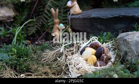 Uova di cioccolato pasquale nel nido nascosto nel giardino con due coniglietti sullo sfondo. Profondità di campo bassa Foto Stock