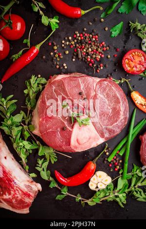 Fusto di vitello crudo (ossobuco), su un fondo di gesso nero con pomodori ciliegini, pepe caldo ed erbe aromatiche. Pronto per la cottura Foto Stock