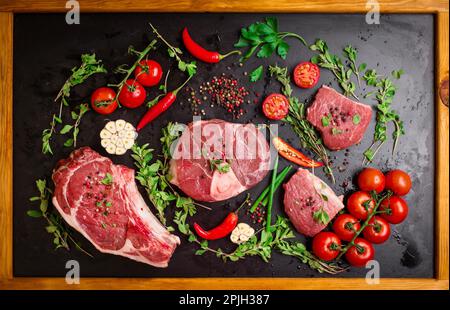 Diversi tipi di bistecche. Carne cruda assortita su un fondo di gesso nero con struttura in legno. Costola occhio sull'osso, gambo di vitello (ossobuco) Foto Stock