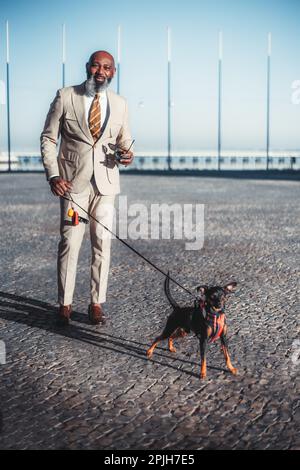 Ripresa verticale di un uomo nero calvo con una barba che cammina il suo cane sul marciapiede di Lisbona. Tiene il guinzaglio in una mano e i suoi occhiali da sole nel Foto Stock