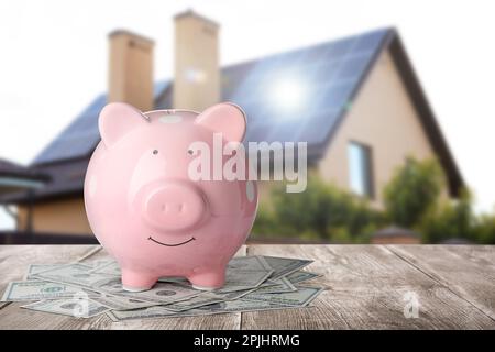 Piggy bank e denaro su superficie di legno e vista sfocata di bella casa, spazio per il testo. Concetto ipotecario Foto Stock