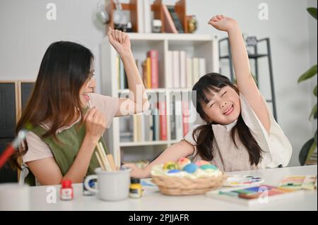 Eccitato e overjoyed giovane ragazza asiatica che alza la mano, ama dipingere uovo di Pasqua per le vacanze con la madre in cucina. Foto Stock