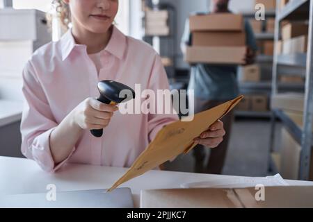 Primo piano della lavoratrice della posta che scansiona il codice sulla busta con lettera di consegna rapida mentre si siede davanti alla fotocamera sul posto di lavoro Foto Stock