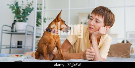La donna matura si trova sul pavimento con il pinscher mini del cane dello sclittino, godendo pomeriggio nel paese, il concetto di animale come membro della famiglia Foto Stock