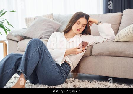 Si siede sul pavimento. Giovane donna in camicia bianca e jeans è a casa Foto Stock
