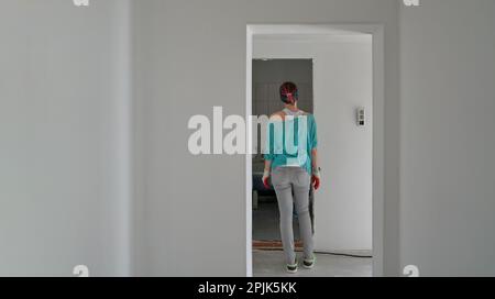 Donna si erge in un corridoio dipinto di fresco e guarda nella stanza. Concetto di riparazione e ristrutturazione della casa. Foto Stock