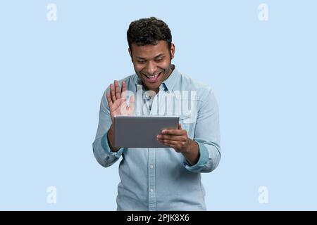 Ritratto di un giovane ragazzo indiano che agita la mano a qualcuno sul suo tablet pc. Isolato su sfondo blu pastello. Foto Stock