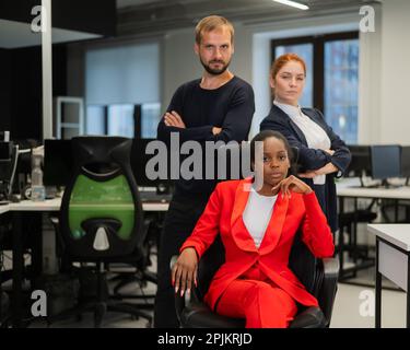 Donna caucasica dai capelli rossi, uomo caucasico bearded in piedi dietro una giovane donna afroamericana seduta in ufficio. Foto Stock