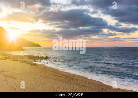Luce del tramonto con colori dorati e rosa che filtrano attraverso le nuvole sopra la silhouette di una costa con in primo piano una spiaggia di sabbia con rocce e. Foto Stock