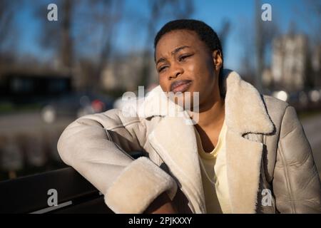 Ritratto all'aperto di una triste donna afro-americana nella giornata di sole. È seduta su panca per strada. Foto Stock