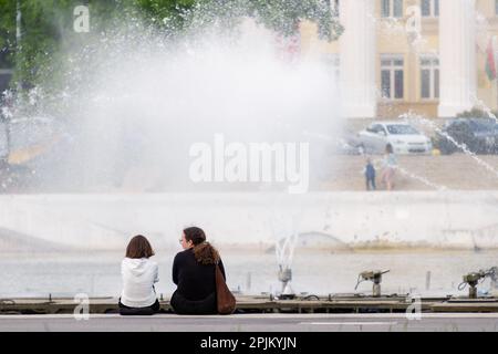 Minsk, Bielorussia - 31 maggio 2022: Le ragazze si siedono sul terrapieno sullo sfondo di una fontana funzionante che spruzzi. Schizzo della città Foto Stock