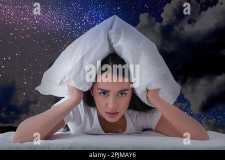Giovane donna infastidita che copre le orecchie con cuscino e bel cielo stellato di notte sullo sfondo Foto Stock