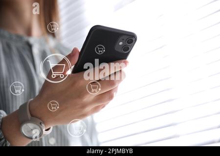 Concetto di consegna. Donna che utilizza un telefono cellulare moderno per lo shopping online al chiuso, primo piano. Illustrazioni del carrello di mercato Foto Stock