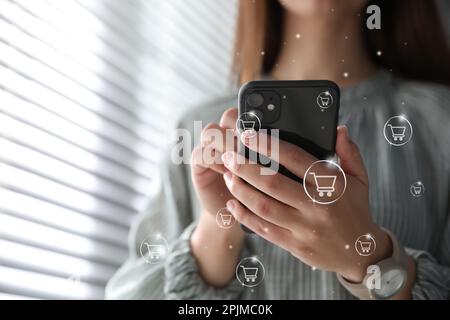 Concetto di consegna. Donna che utilizza un telefono cellulare moderno per lo shopping online al chiuso, primo piano. Illustrazioni del carrello di mercato Foto Stock