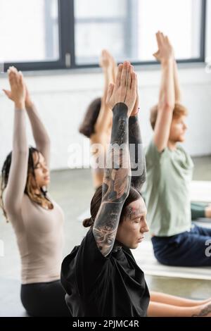 Tatuato uomo che fa mudra anjali mentre si siede vicino a persone sfocate in classe yoga, immagine stock Foto Stock