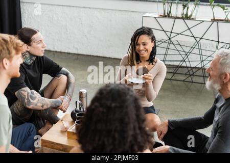 donna afro-americana felice con dreadlocks che tiene il tè del puer compresso vicino alla gente in studio di yoga, immagine di scorta Foto Stock