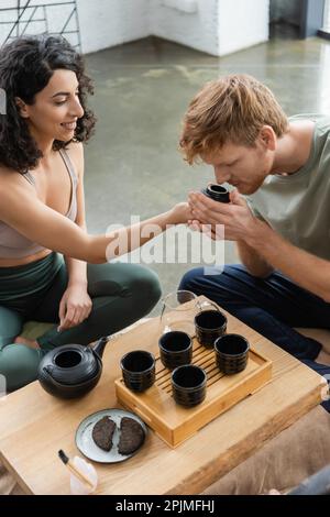 curly medio orientale donna sorridente mentre dando per odorare tè puer a redhead uomo, immagine stock Foto Stock