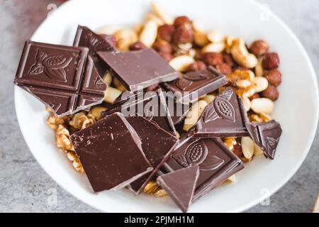 Cioccolato con frutta secca, anacardi, noci, nocciole in un piatto bianco sul tavolo, vista dall'alto, primo piano Foto Stock