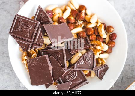 Cioccolato con frutta secca, anacardi, noci, nocciole in un piatto bianco sul tavolo, vista dall'alto, primo piano Foto Stock