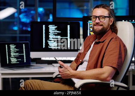 Giovane uomo sorridente INGEGNERE IT in occhiali utilizzando smartphone e guardando la fotocamera mentre si siede in poltrona accanto al suo posto di lavoro Foto Stock