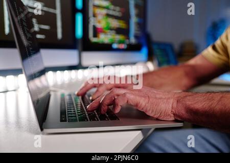 Concentratevi sulle mani dei programmatori maschi anziani che scrivono sulla tastiera del laptop mentre decodificano i dati sullo schermo in un ufficio buio a tarda notte Foto Stock