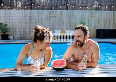 Coppia felice che gode il tempo di estate in piscina, con le bevande e l'anguria. Foto Stock