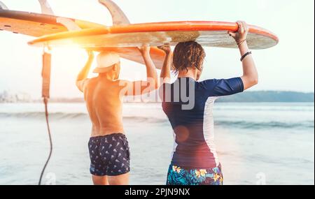 Ragazzo con suo padre con tavole da surf sulla testa che cammina nelle onde sulla spiaggia di Udawalawe in Sri Lanka. Vacanza attiva in famiglia o sport attivo pe Foto Stock