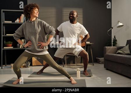 Felice giovane donna attiva e suo marito che fa esercizi sui tappetini durante l'allenamento mattutino nel soggiorno a piacere o nel fine settimana Foto Stock