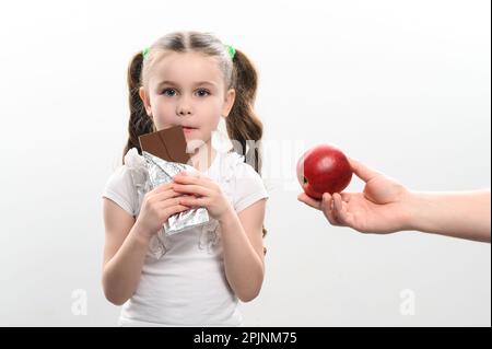 Ritratto di una bambina su sfondo bianco, sceglie il cioccolato su una mela, cibo sano vs cibo spazzatura. Foto Stock