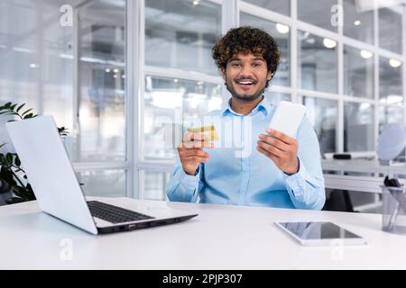 Felice giovane uomo seduto in ufficio alla scrivania con telefono e carta di credito. Effettua acquisti online, paga fatture. Sorridendo guardando la fotocamera. Foto Stock