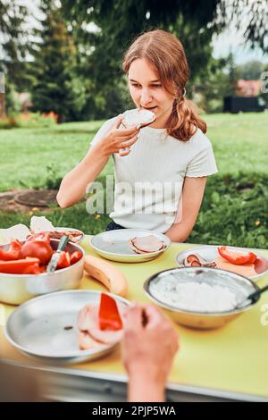 La famiglia fa colazione all'aperto in campeggio durante le vacanze estive. Pane, cottage cheese, carne fredda, pomodori, frutta e tazze di caffè sul tavolo Foto Stock