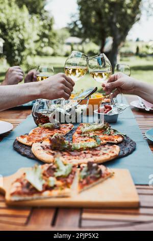 Gli amici fanno brindisi durante il picnic estivo cena all'aperto in un giardino di casa. Primo piano di persone che tengono bicchieri da vino con vino bianco sul tavolo con piz Foto Stock