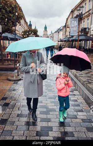 Madre e sua bambina che tengono gli ombrelloni rosa e blu che camminano in un centro in una notte d'autunno piovosa e cupa Foto Stock