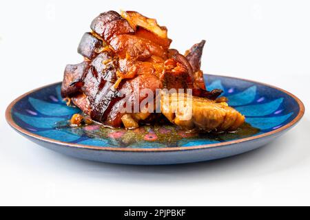 Pancia di maiale al forno e aglio su piatto blu decorativo, spicchio su sfondo bianco. Foto Stock