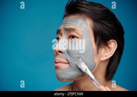 Faccia di giovane uomo che applica la maschera idratante del viso con il pennello Foto Stock
