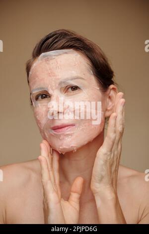 Donna sorridente di mezza età che applica maschera anti-rughe foglio sul viso Foto Stock